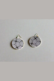 Pair of black and white patterned earrings on a green surface with feet wearing sandals in the foreground.