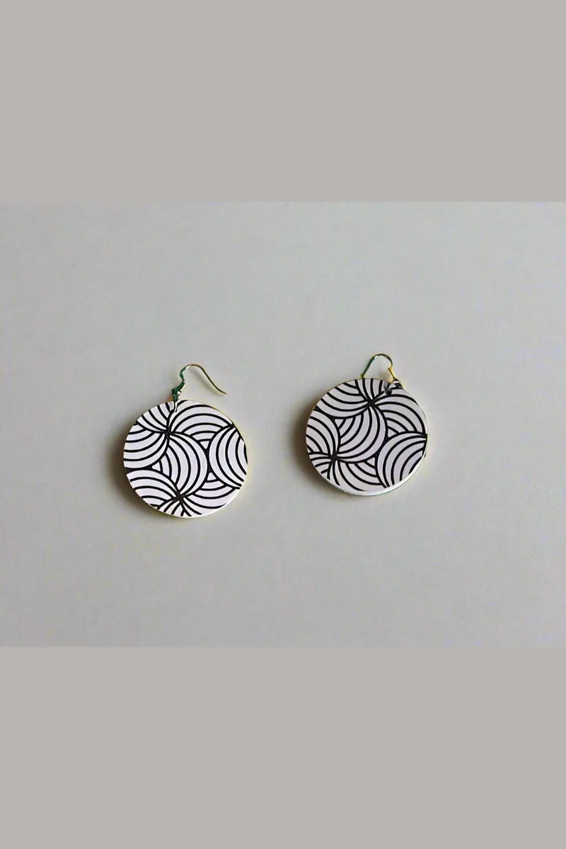 Pair of black and white patterned earrings on a green surface with feet wearing sandals in the foreground.