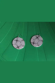 Pair of black and white patterned earrings on a green surface with feet wearing sandals in the foreground.