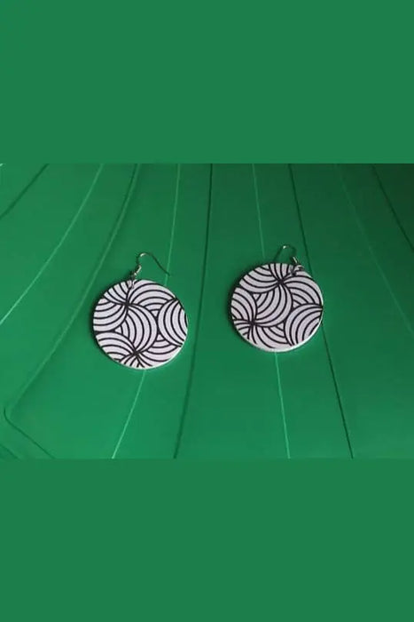 Pair of black and white patterned earrings on a green surface with feet wearing sandals in the foreground.